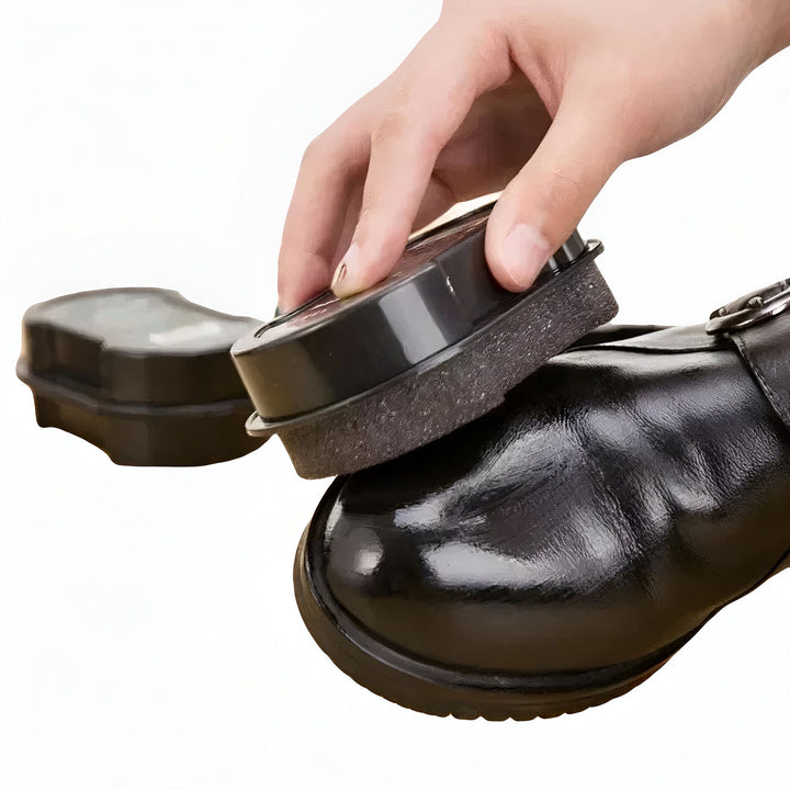 Une main polit une chaussure en cuir noir avec un applicateur de cirage noir. On voit un deuxième applicateur de cirage en arrière-plan., 45286736593077