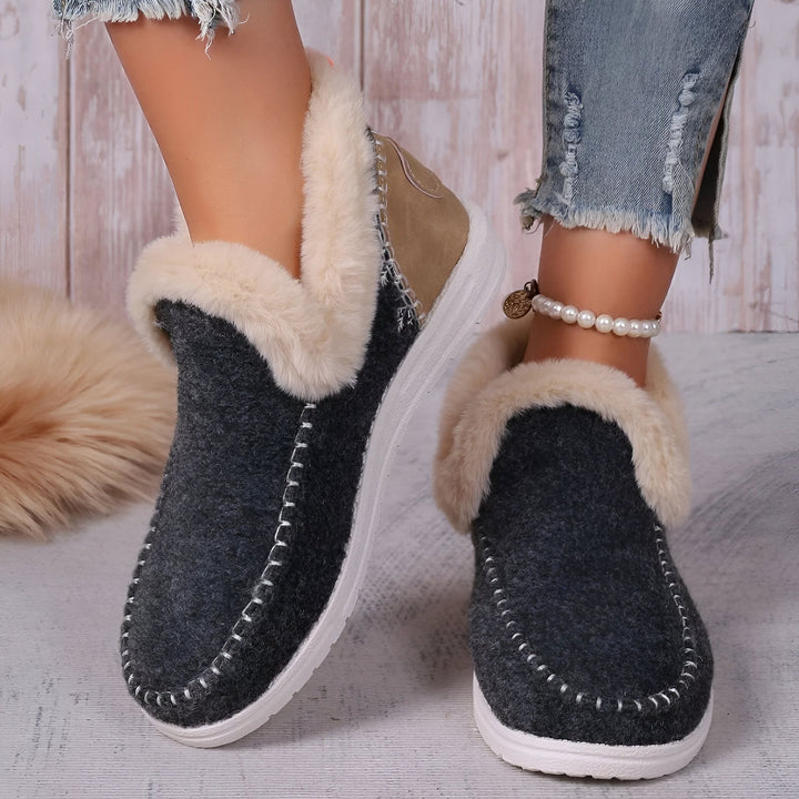 Gros plan de chaussons d'intérieur gris avec bordure en fausse fourrure beige, portés par une femme avec un jean effiloché et un bracelet de perles à la cheville. Chaussures confortables, idéales pour la maison., 45286658375861