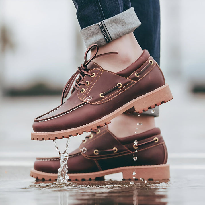 Gros plan sur des chaussures bateau en cuir marron portées dans une flaque d'eau. Les chaussures sont lacées et ont une semelle épaisse. Une personne en jean bleu est debout., 45287041466549,45287041499317,45287041532085,45287041564853,45287041597621,45287041630389,45287041663157,45287041695925,45287041728693,45287041761461