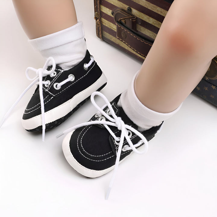 Gros plan de pieds de bébé portant des baskets noires et blanches avec des lacets non attachés. Une valise vintage est visible en arrière-plan., 45286825787573,45286825820341,45286825853109,45286825885877,45286825918645,45286825951413,45286825984181,45286826016949,45286826049717,45286826082485,45286826115253,45286826148021