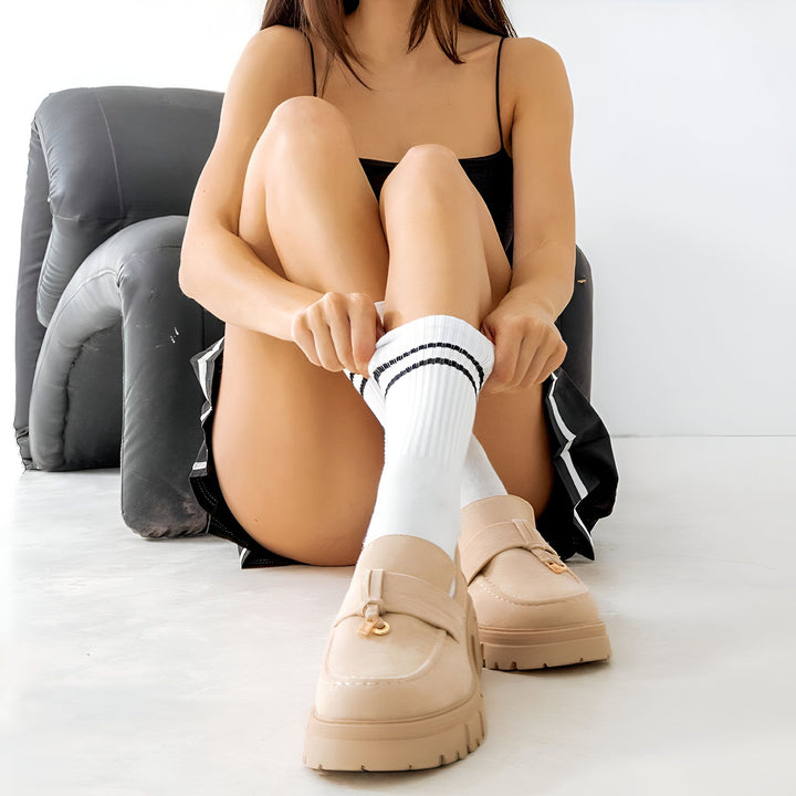Une femme assise tire sur ses chaussettes blanches à rayures noires, portant des mocassins beige. Elle est vêtue d'un débardeur noir et d'un short noir et blanc, assise devant une chaise noire gonflable., 45286817628341,45286817661109,45286817693877,45286817726645,45286817759413,45286817792181
