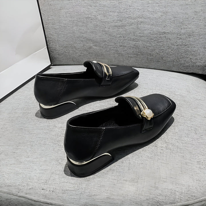 Deux mocassins noirs en cuir sur un coussin gris. Les chaussures sont ornées d'une boucle dorée et d'une perle. Élégance et confort pour toutes les occasions., 45287056277685,45287056310453,45287056343221,45287056375989,45287056408757,45287056441525,45287056474293,45287056507061,45287056539829,45287056572597,45287056605365,45287056638133