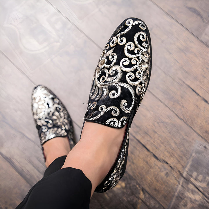 Vue de dessus des pieds portant des mocassins ornementaux. Un mocassin est noir avec des broderies argentées, tandis que l'autre mocassin est argenté avec des broderies noires. Portés avec un pantalon noir sur un plancher de bois., 45286994706613,45286994739381,45286994772149,45286994804917,45286994837685,45286994870453,45286994903221,45286994935989,45286994968757,45286995001525,45286995034293,45286995067061,45286995099829,45286995132597,45286995165365,45286995198133