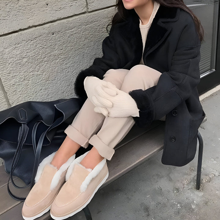 Une femme assise sur un banc, portant un manteau noir bordé de fourrure, un pantalon beige, des chaussons beiges fourrés et des gants en laine écrus. Un sac en cuir noir est à côté d'elle., 45286653395125,45286653427893,45286653460661,45286653493429,45286653526197,45286653558965,45286653591733,45286653624501,45286653657269,45286653690037,45286653722805,45286653755573,45286653788341,45286653821109,45286653853877,45286653886645,45286653919413,45286653952181,45286653984949,45286654017717,45286654050485