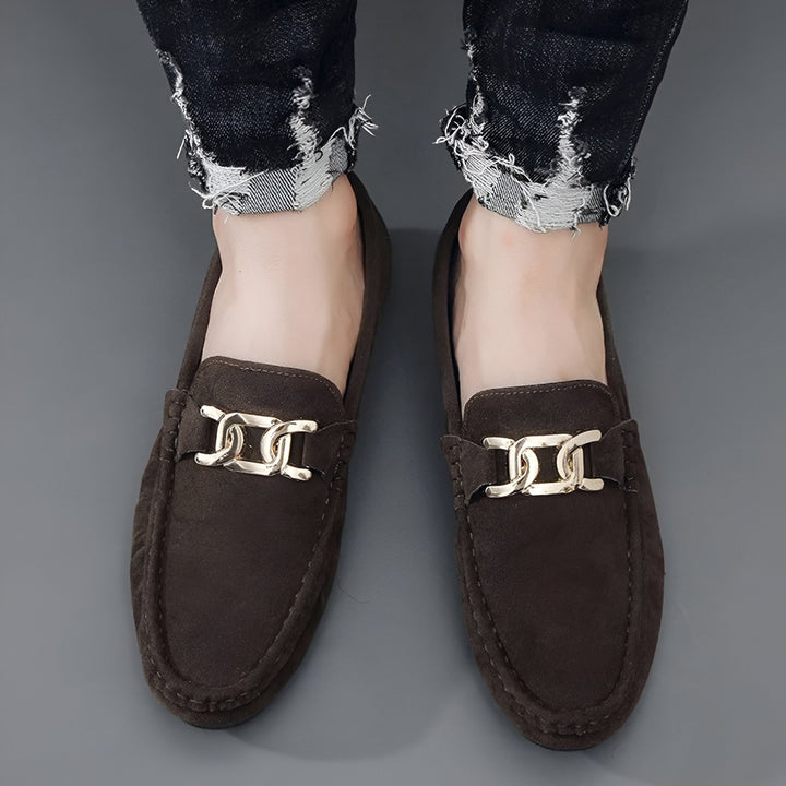 Vue de dessus d'une personne portant des mocassins marron en daim avec une chaîne décorative dorée, associés à un jean noir délavé et effiloché., 45286663127221,45286663159989,45286663192757,45286663225525,45286663258293,45286663291061,45286663323829,45286663356597,45286663389365,45286663422133,45286663454901,45286663487669,45286663520437,45286663553205,45286663585973,45286663618741,45286663651509,45286663684277,45286663717045,45286663749813,45286663782581,45286663815349,45286663848117,45286663880