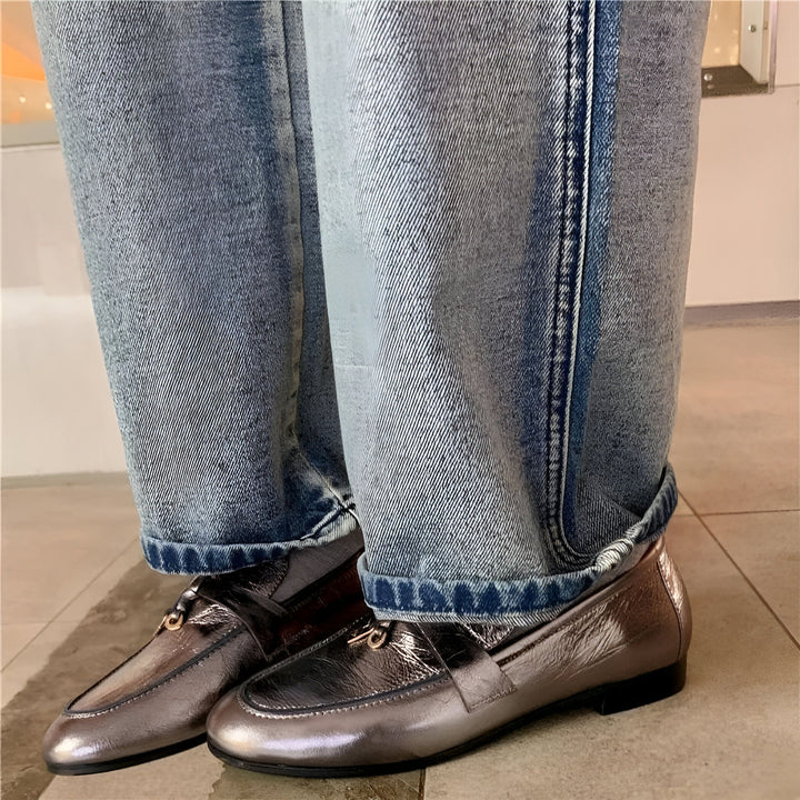 Vue rapprochée d'une personne portant un jean bleu ample retroussé et des mocassins argentés avec une bride et une boucle métallique. Le sol est carrelé de couleur neutre. Mode décontractée et élégante., 45286733185205,45286733217973,45286733250741,45286733283509,45286733316277,45286733349045,45286733381813