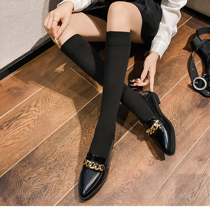 Femme assise portant des mocassins noirs vernis avec chaîne dorée et des chaussettes montantes noires, sur un sol en bois. Détail d'une tenue à la mode., 45286940147893,45286940180661,45286940213429,45286940246197,45286940278965,45286940311733,45286940344501,45286940377269,45286940410037