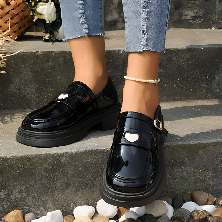 Gros plan sur une femme portant des chaussures vernies noires avec un détail en forme de cœur blanc et un jean bleu délavé. Elle porte une chevillère en perles., 45286866419893,45286866452661,45286866485429,45286866518197,45286866550965,45286866583733