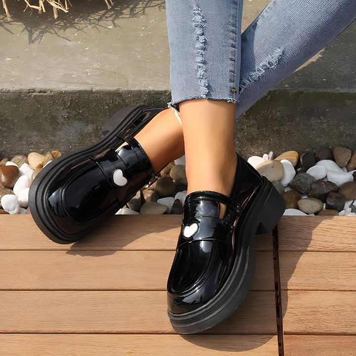 Gros plan de mocassins vernis noirs avec un motif de cœur blanc portés avec un jean bleu délavé. Les chaussures sont sur une surface en bois, avec des galets et une bordure en béton en arrière-plan., 45286866419893,45286866452661,45286866485429,45286866518197,45286866550965,45286866583733