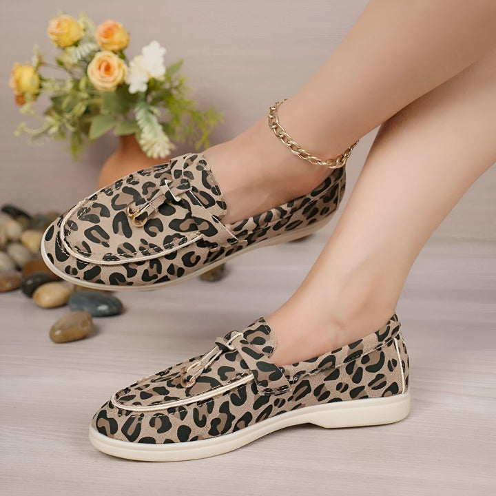 Chaussures mocassins léopard portées par une femme avec une chaîne de cheville dorée. Décor floral et galets en arrière-plan. Style décontracté et tendance., 45286648250549,45286648283317,45286648316085,45286648348853,45286648381621,45286648414389,45286648447157,45286648479925