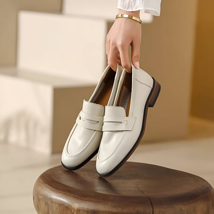 Gros plan de mocassins en cuir blanc crème, tenus par une main avec un bracelet doré, sur un tabouret en bois., 45286733414581,45286733447349,45286733480117,45286733512885,45286733545653,45286733578421,45286733611189