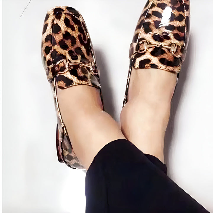 Vue rapprochée de pieds portant des mocassins en cuir verni à motif léopard, avec une boucle dorée, portés avec un pantalon noir, sur un fond blanc., 45287153991861,45287154024629,45287154057397,45287154090165,45287154122933,45287154155701