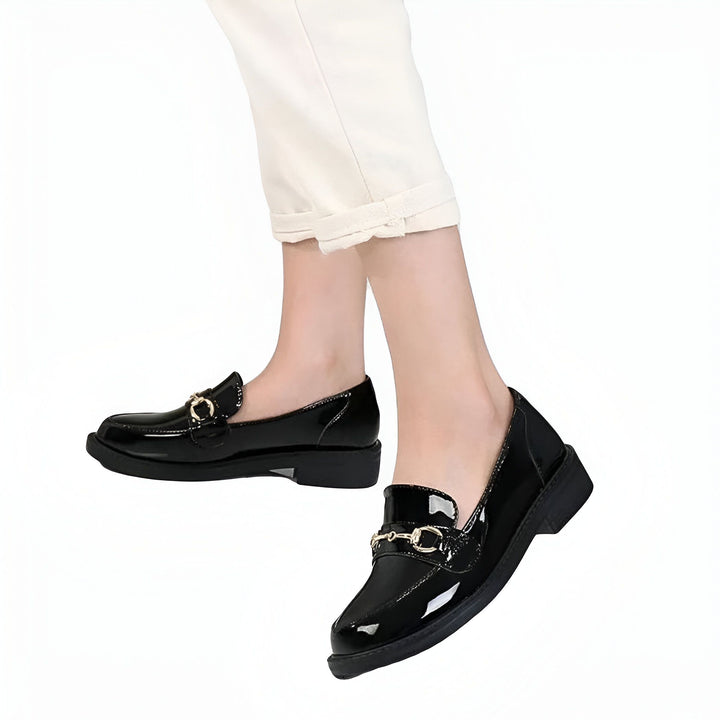 Gros plan de mocassins vernis noirs avec ornement métallique portés avec un pantalon blanc. Chaussures élégantes pour femmes., 45286727549109,45286727581877,45286727614645,45286727647413,45286727680181,45286727712949,45286727745717,45286727778485,45286727811253