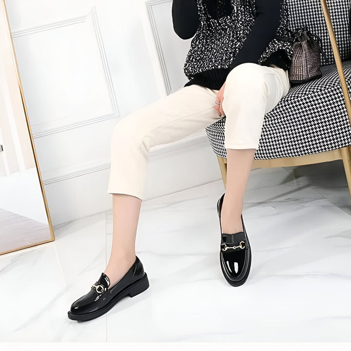 Femme assise portant des mocassins vernis noirs avec des détails dorés, associée à un pantalon beige et un pull. Un sac à main et un fauteuil à motif pied-de-poule complètent le look., 45286727549109,45286727581877,45286727614645,45286727647413,45286727680181,45286727712949,45286727745717,45286727778485,45286727811253