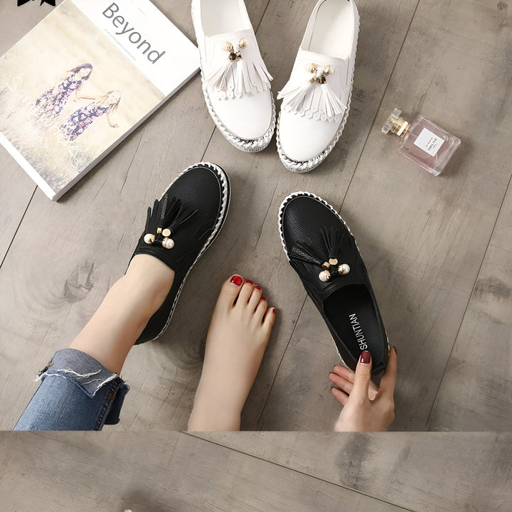 Vue rapprochée de chaussures plates à enfiler, blanches et noires, avec des glands et ornements dorés. Un pied porte une chaussure noire. Un livre, et un parfum complètent la scène., 45286766313653,45286766346421,45286766379189,45286766411957,45286766444725,45286766477493,45286766510261,45286766543029,45286766575797,45286766608565,45286766641333,45286766674101