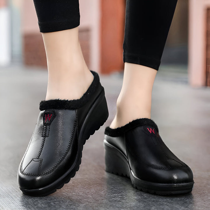 Gros plan sur des pieds portant des chaussons d'intérieur noirs avec une bordure en fausse fourrure. Les chaussons ont un petit talon compensé et un logo "W" rouge brodé., 45286906331317,45286906364085,45286906396853,45286906429621,45286906462389,45286906495157,45286906527925,45286906560693,45286906593461,45286906626229,45286906658997,45286906691765,45286906724533,45286906757301