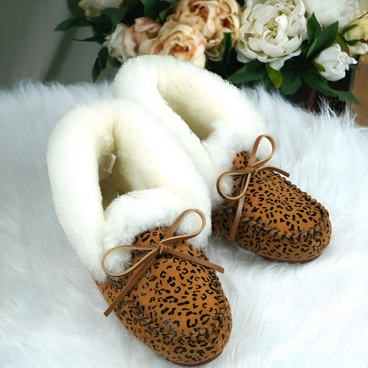Image de chaussons mocassins en peau de mouton à imprimé léopard avec des nœuds en cuir sur un tapis en fausse fourrure, avec des fleurs en arrière-plan. Chaussons doux, confortables et élégants pour femmes., 45286951452853,45286951485621,45286951518389,45286951551157,45286951583925,45286951616693,45286951649461,45286951682229,45286951714997,45286951747765,45286951780533,45286951813301,45286951846069,45286951878837,45286951911605,45286951944373,45286951977141,45286952009909,45286952042677,452869520