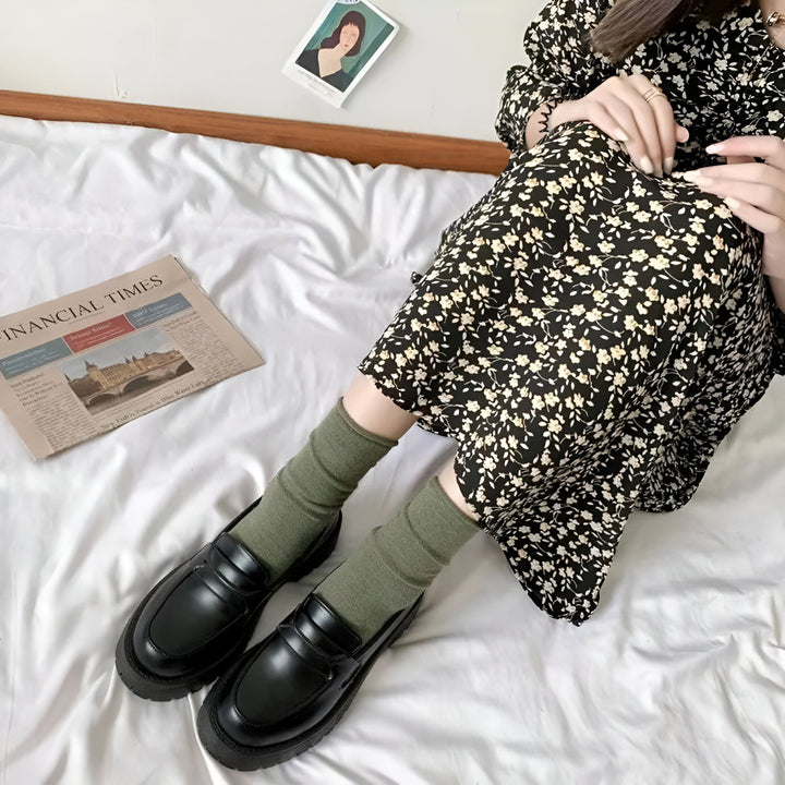 Une femme assise sur un lit blanc, portant une robe fleurie noire, des chaussettes vertes et des mocassins noirs. Un journal "Financial Times" et une petite image sont sur le lit. Style décontracté et confortable., 45286720700597,45286720733365,45286720766133,45286720798901,45286720831669,45286720864437,45286720897205,45286720929973,45286720962741