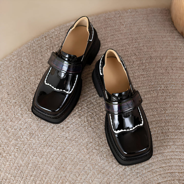 Vue rapprochée de deux chaussures de style écolière noires vernies avec une bride à boucle sur un tapis texturé beige. Détails festonnés blancs., 45286832013493,45286832046261,45286832079029,45286832111797,45286832144565,45286832177333,45286832210101