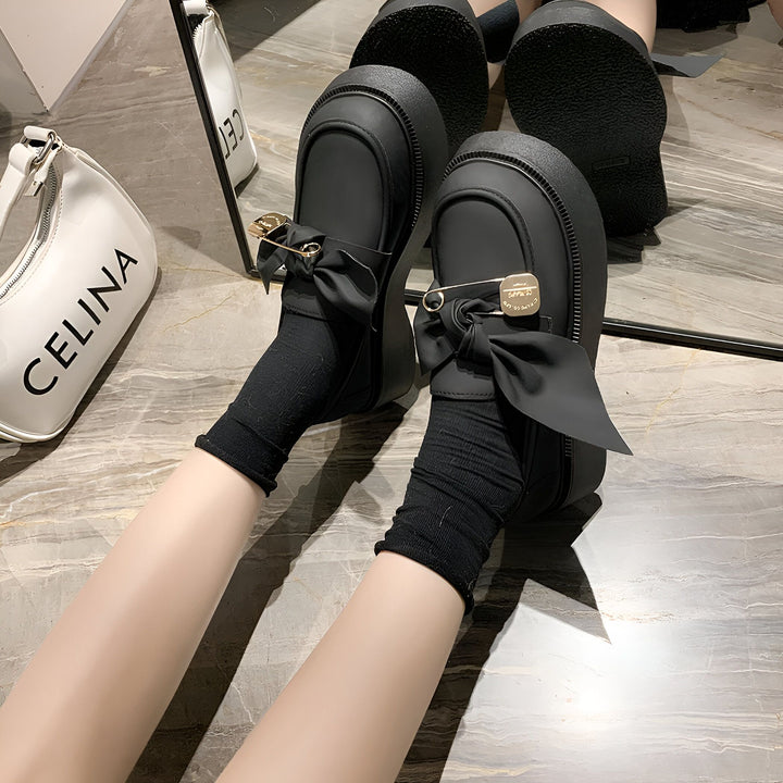 Personne portant des mocassins noirs à plateforme avec des chaussettes noires. Les chaussures sont décorées de nœuds noirs et d'épingles à nourrice dorées. Un sac à main blanc "CELINA" est visible en arrière-plan., 45286885359797,45286885392565,45286885425333,45286885458101,45286885490869,45286885523637,45286885556405,45286885589173,45286885621941,45286885654709,45286885687477,45286885720245