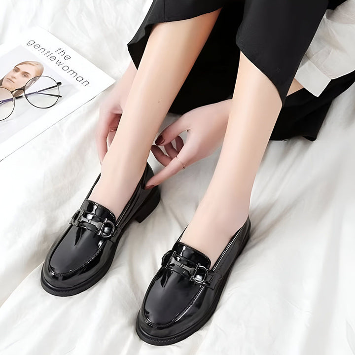 Chaussures mocassins vernies noires avec boucle, portées par une femme avec un pantalon noir. Un magazine et des lunettes sont à côté sur un drap blanc., 45286626295989,45286626328757,45286626361525,45286626394293,45286626427061