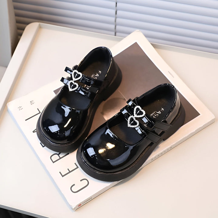 Paire de chaussures vernies noires pour filles, avec brides à boucles en forme de cœur strass, posées sur un livre blanc sur une table claire. Chaussures élégantes et mignonnes pour enfants., 45286682820789,45286682853557,45286682886325,45286682919093,45286682951861,45286682984629,45286683017397,45286683050165,45286683082933,45286683115701,45286683148469,45286683181237,45286683214005,45286683246773,45286683279541,45286683312309,45286683345077,45286683377845,45286683410613,45286683443381,4528668347614