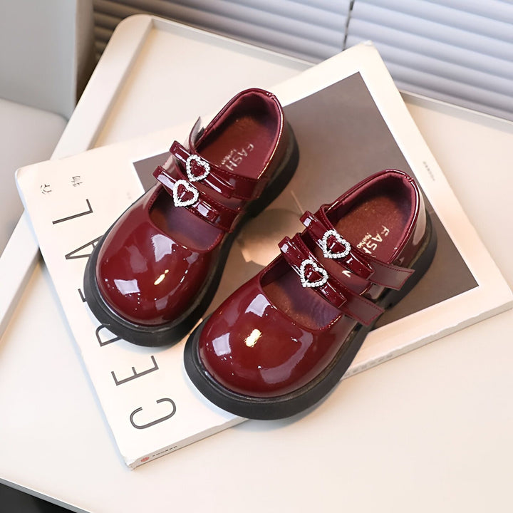 Deux chaussures vernies bordeaux pour enfant avec brides ornées de cœurs brillants, reposant sur un magazine. Chaussures élégantes et brillantes pour petite fille., 45286682820789,45286682853557,45286682886325,45286682919093,45286682951861,45286682984629,45286683017397,45286683050165,45286683082933,45286683115701,45286683148469,45286683181237,45286683214005,45286683246773,45286683279541,45286683312309,45286683345077,45286683377845,45286683410613,45286683443381,45286683476149,45286683508917