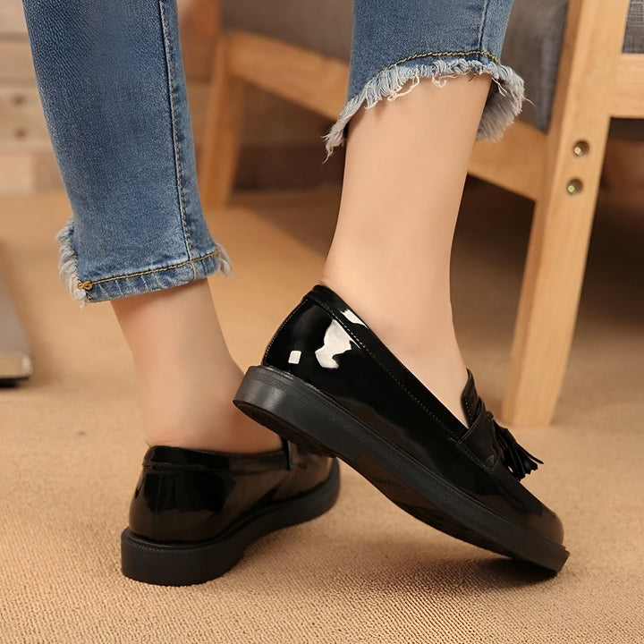 Une femme portant des mocassins noirs vernis avec des glands et un jean effiloché au niveau des chevilles. La photo met en valeur les chaussures et le style vestimentaire., 45286638452917,45286638485685,45286638518453,45286638551221,45286638583989,45286638616757