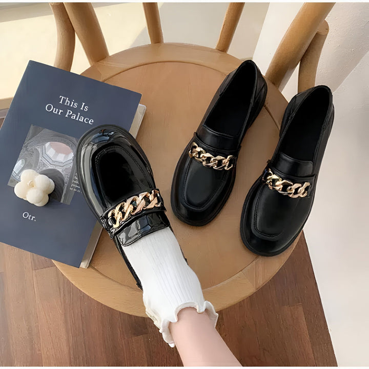 Trois mocassins noirs avec chaînes dorées sur une chaise en bois, à côté d'un livre intitulé "This Is Our Palace". Une personne porte une chaussette blanche et un mocassin. Chaussures élégantes et tendance., 45287065419957,45287065452725,45287065485493,45287065518261,45287065551029,45287065583797